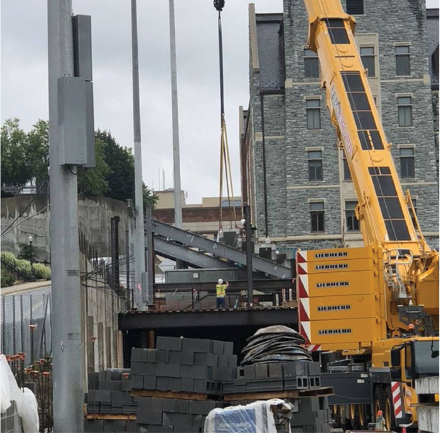 Construction site with crane lifting steel beams; a worker stands below.
