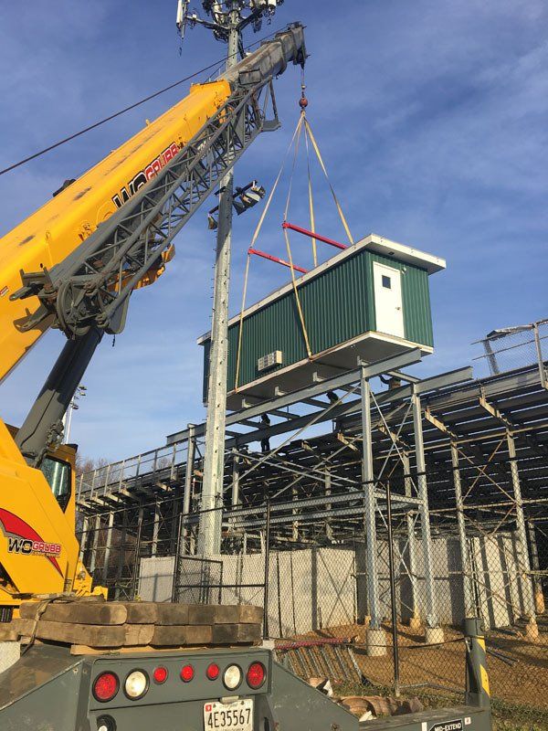 A crane lifts a green utility shed to place it atop stadium scaffolding near light poles.