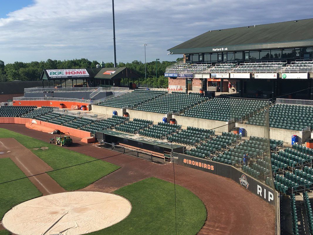 Baseball stadium interior view: green field, brown dirt, empty green seats, orange accents, overcast sky.
