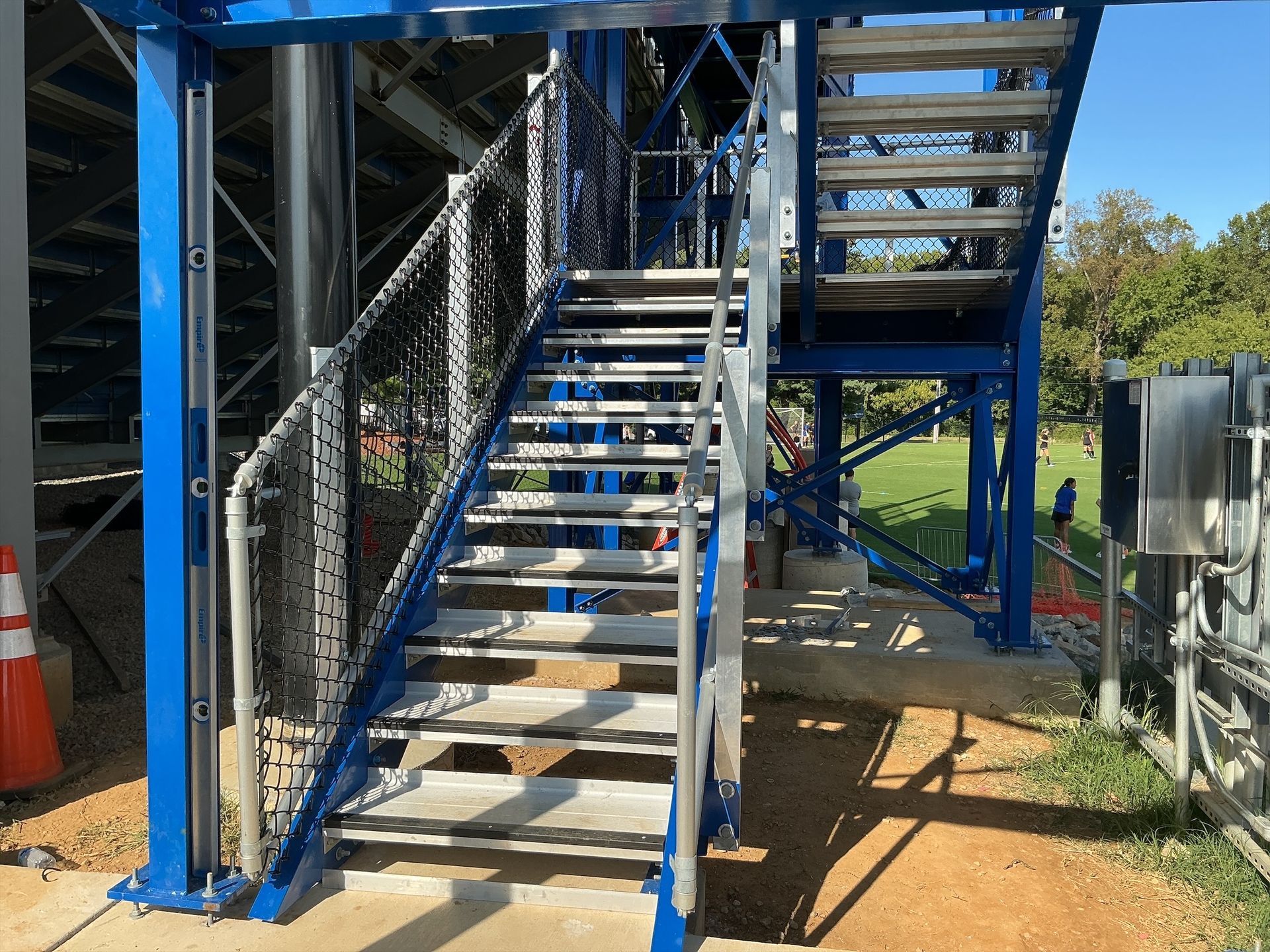 Metal staircase with blue supports, leading up to bleachers. Black netting on the left side.