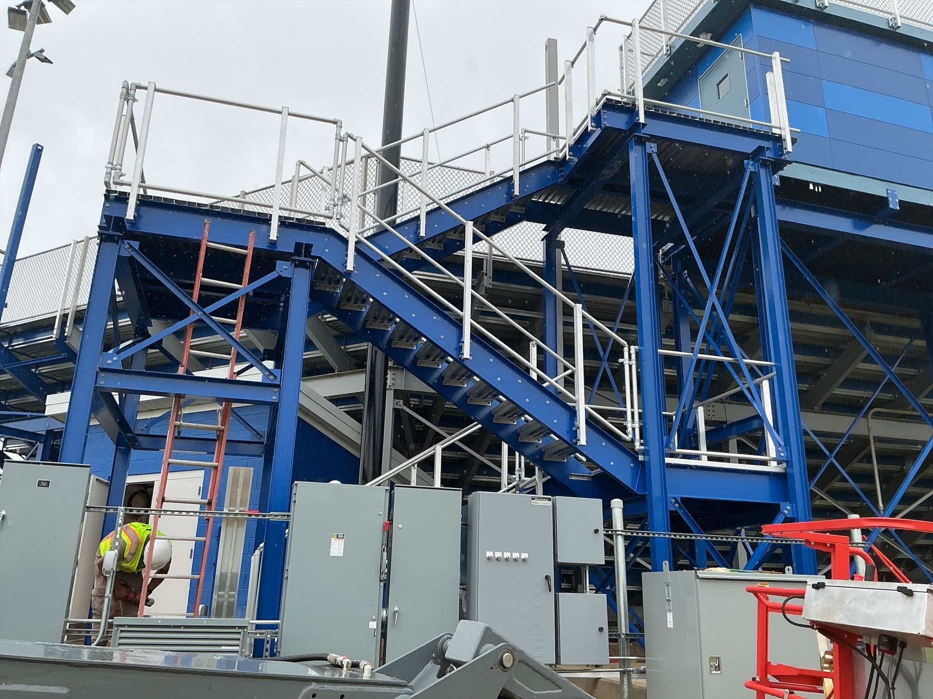 Blue metal staircase and platform with railings; a worker in a safety vest stands nearby.