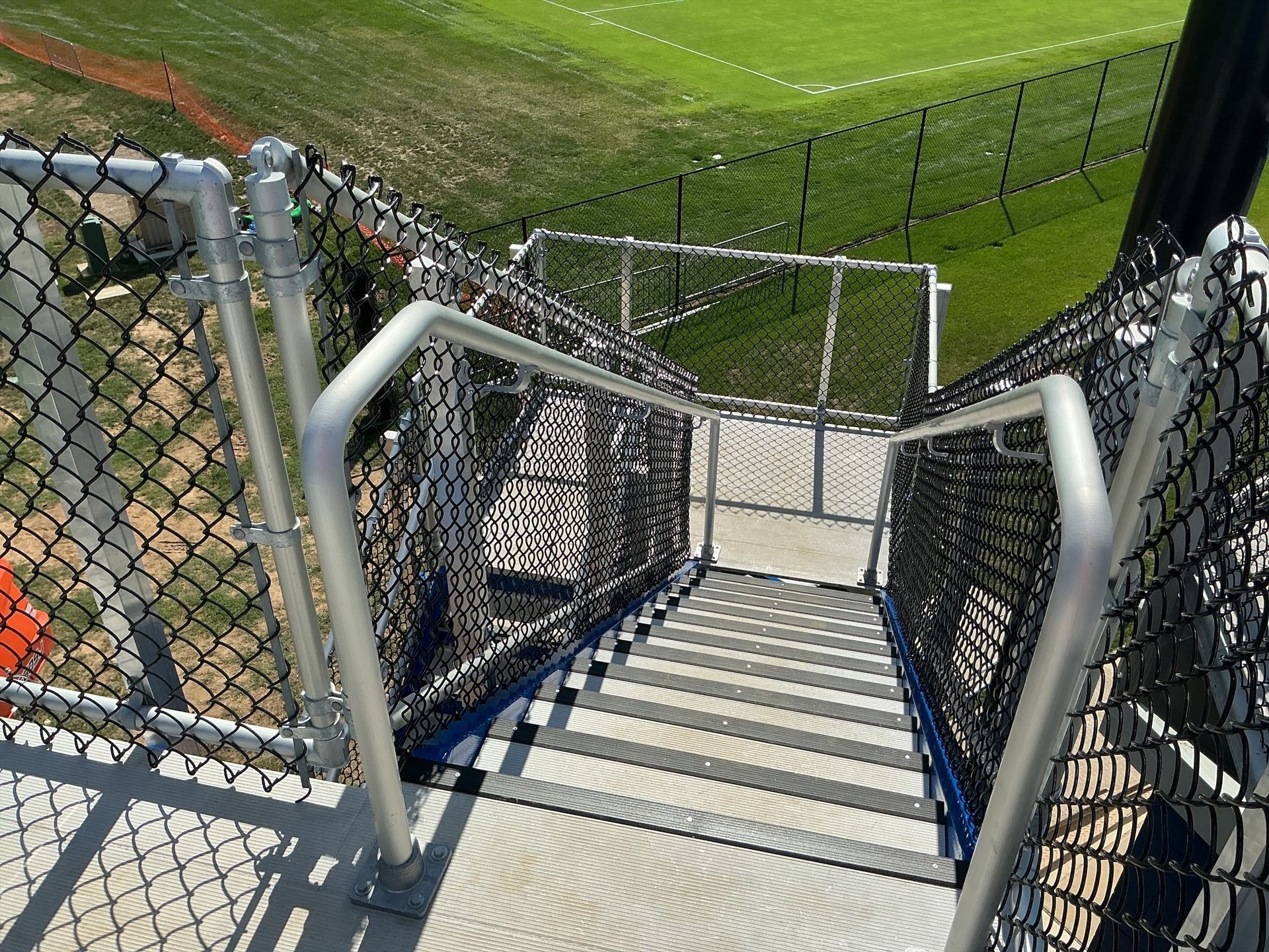 Staircase descending from a raised platform, with metal handrails and chain-link fencing, overlooking a grassy field.