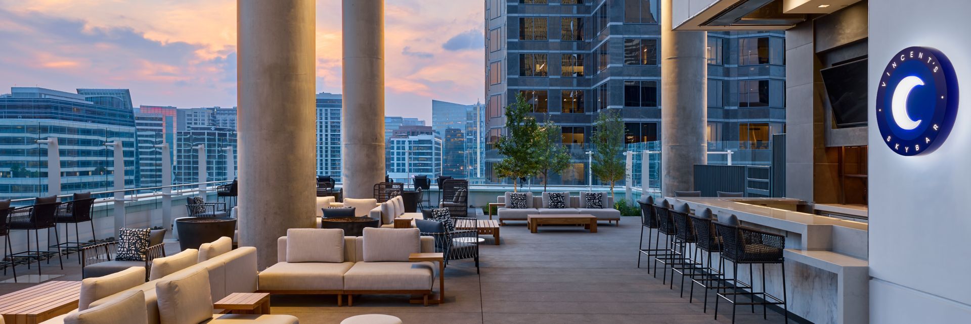 Rooftop bar with city views. Beige seating, bar stools, and a blue moon sign. Cloudy sky at dusk.