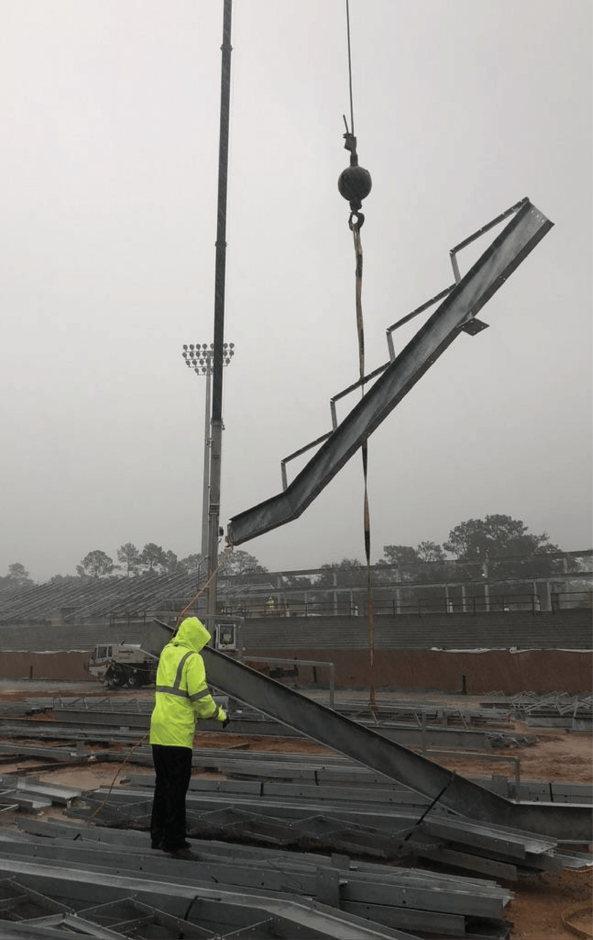 Construction worker in a yellow vest guides a steel staircase being lifted by a crane on a foggy construction site.