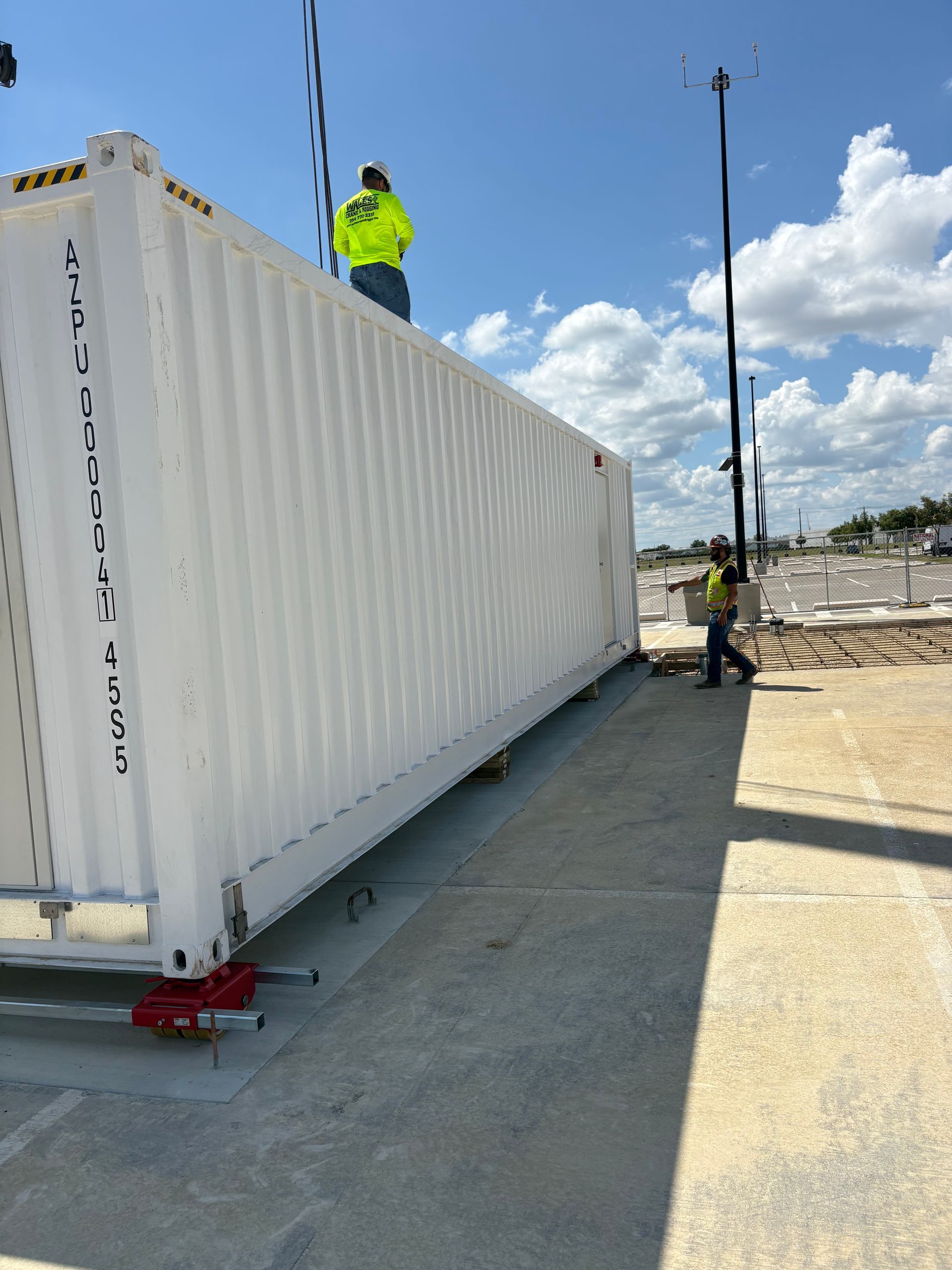Two workers lift a white shipping container. One stands on top, the other on the ground. Bright sunlight.