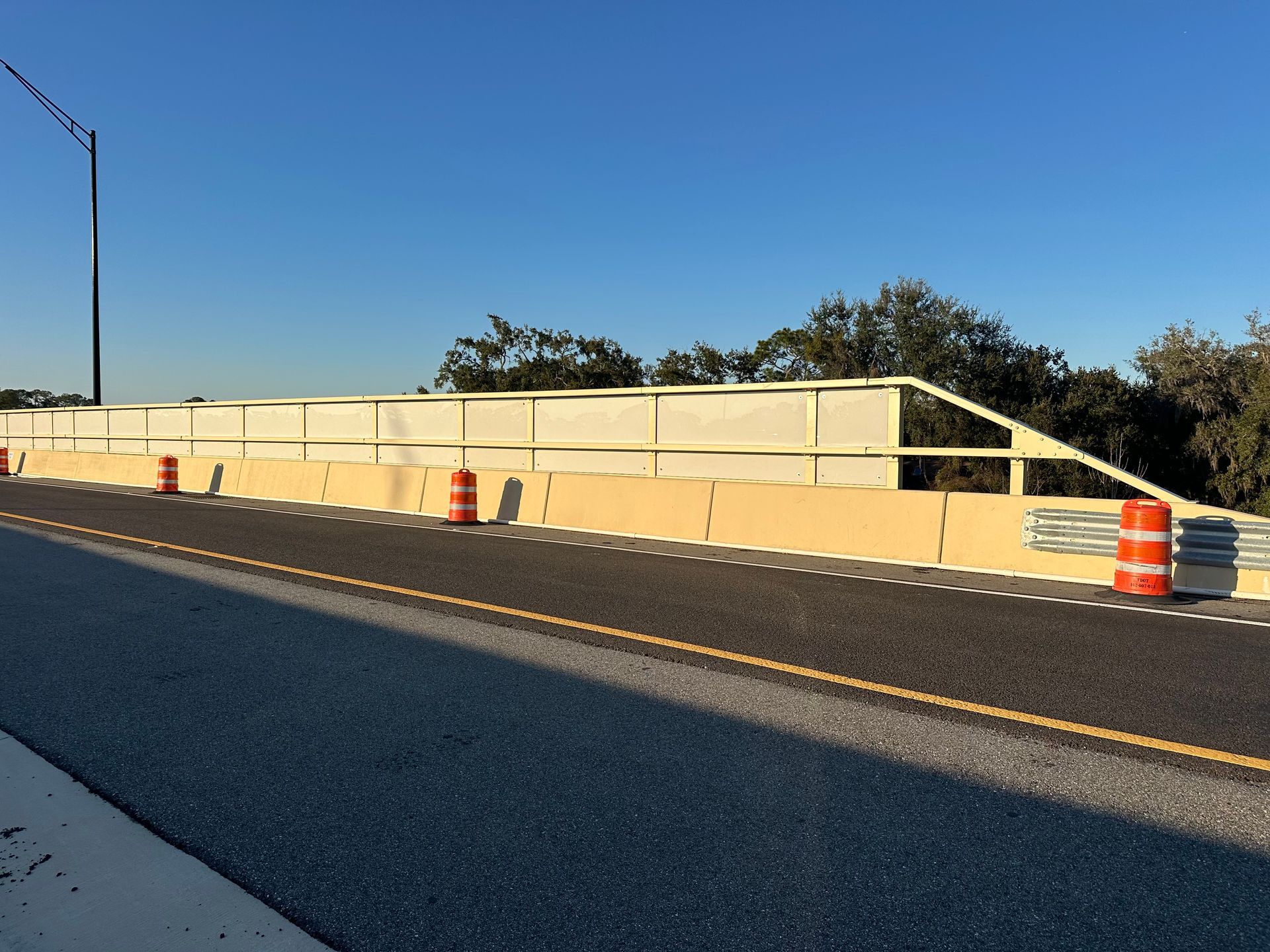 Roadside view of a bridge with jersey barriers, fencing, and orange cones under a clear blue sky.