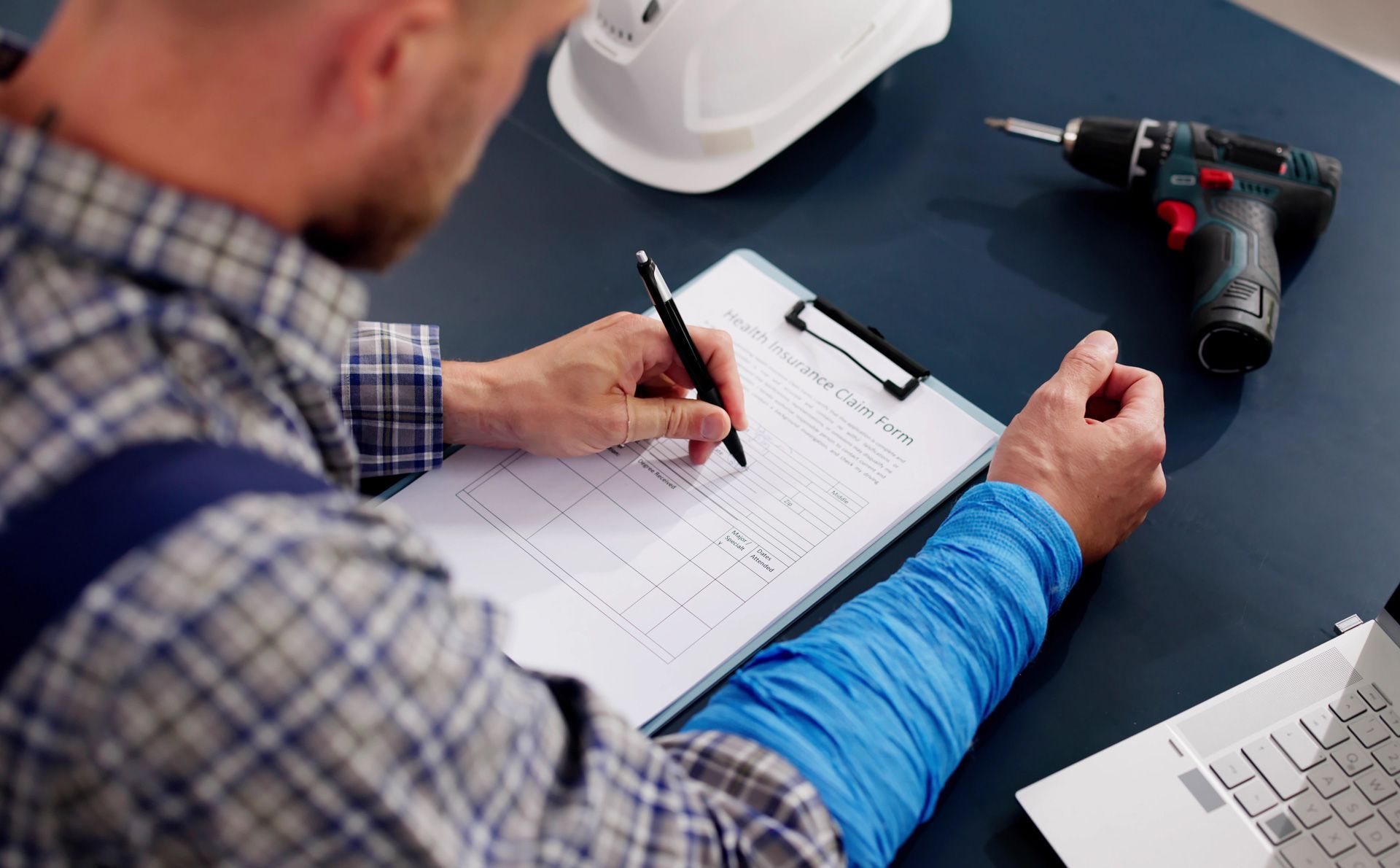 Construction worker in plaid shirt and blue wrap, writing on clipboard at desk with tools and helmet.