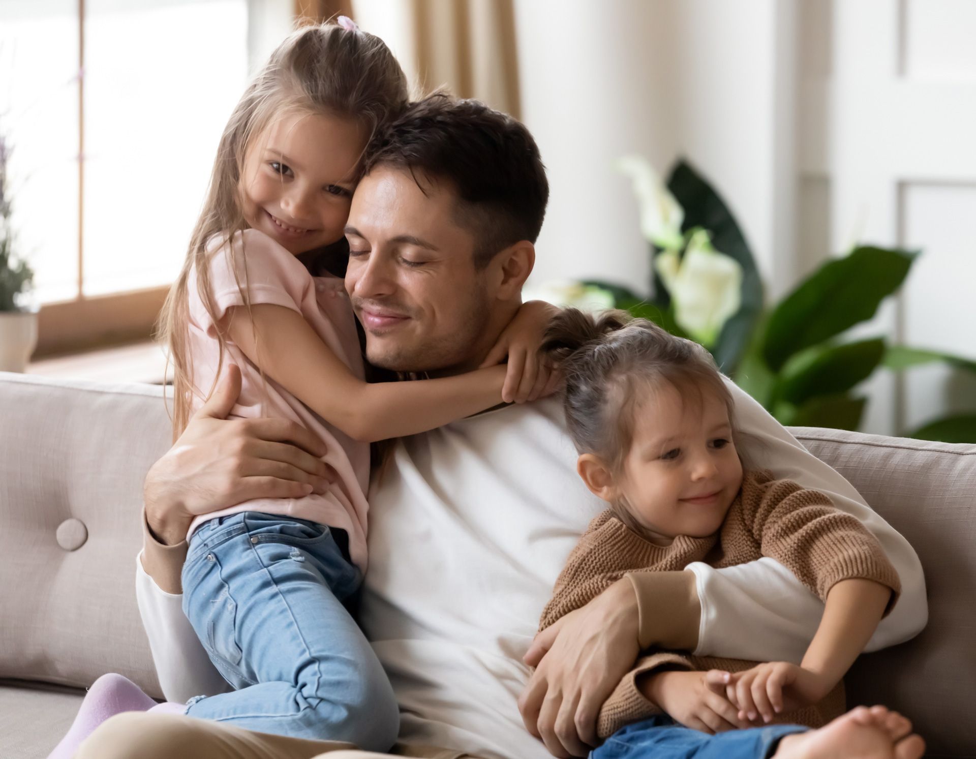 Father sitting on couch, hugged by two young daughters; smiles in a bright living room.
