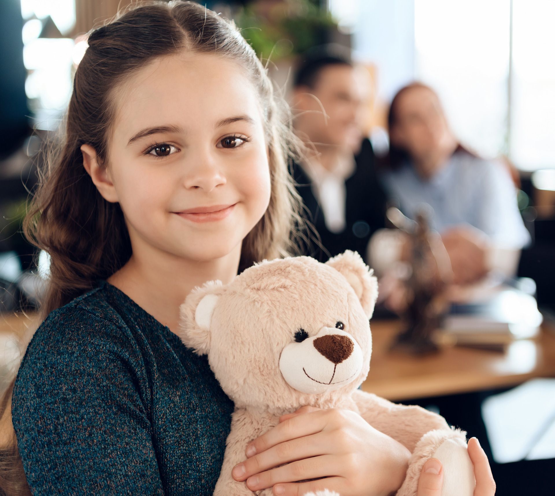 Girl holding a teddy bear, smiling. Family in background at table.