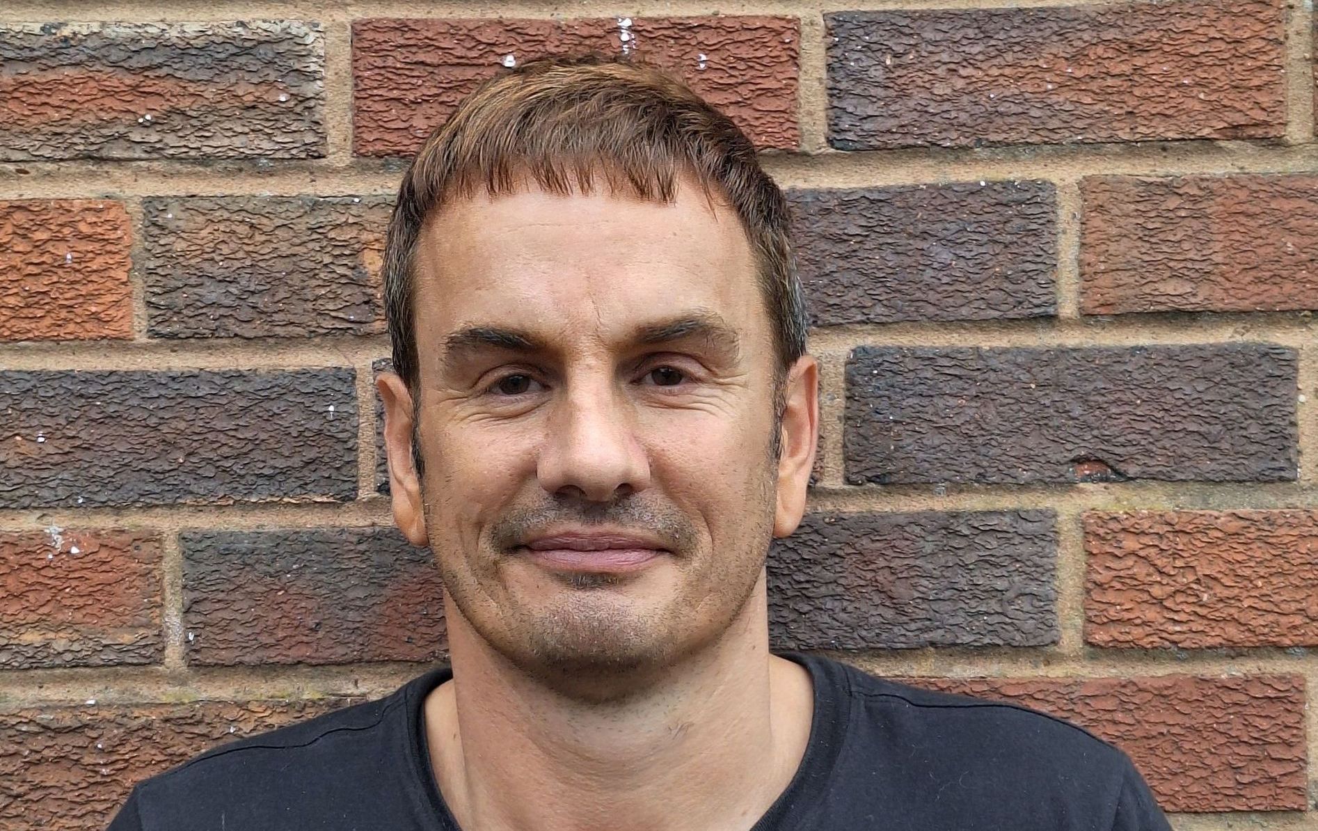 Man with short, dark hair in front of a brick wall, wearing a black shirt, looking at the camera.