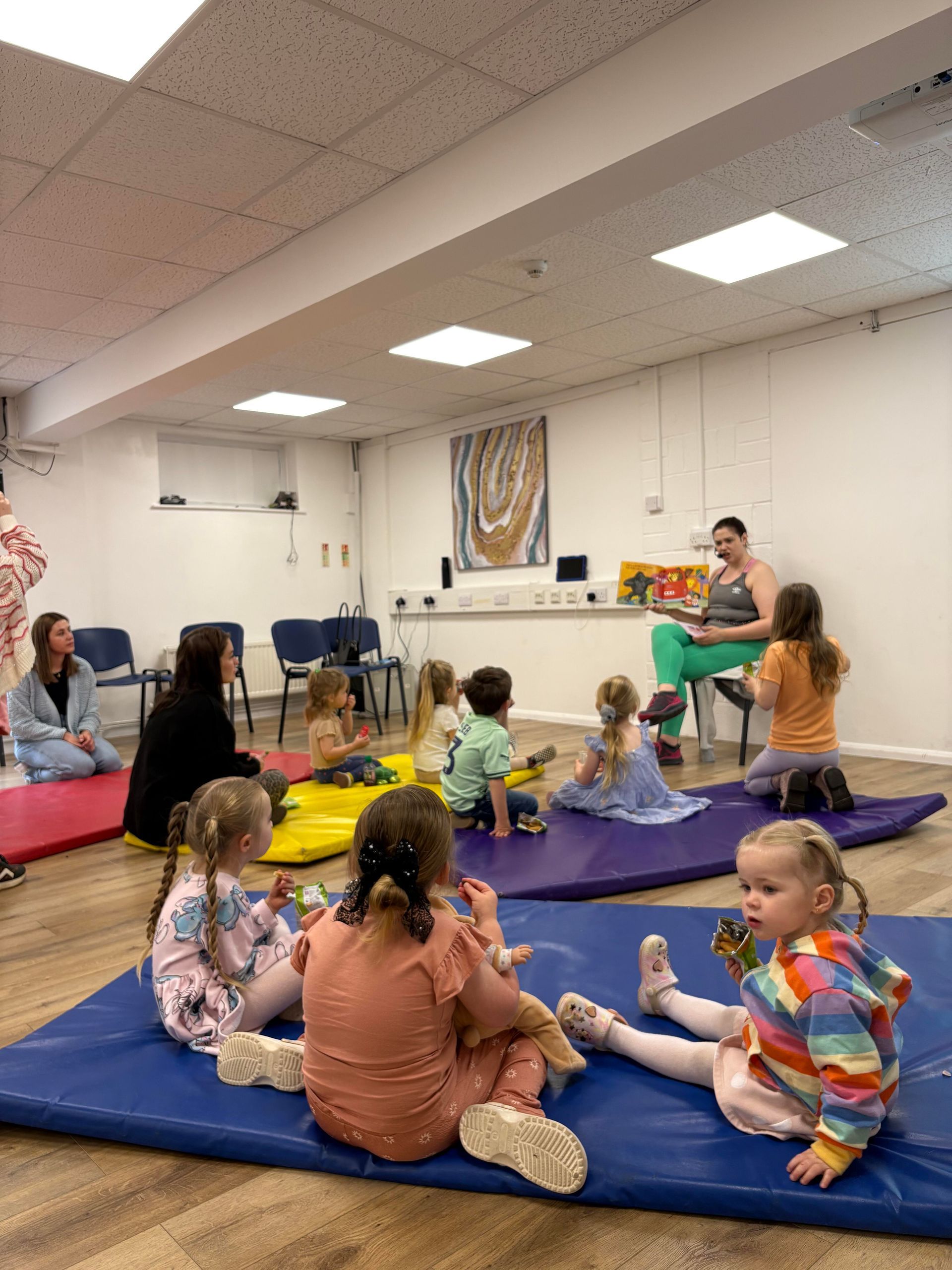 Children sit on mats as an adult reads a book in a brightly lit room.