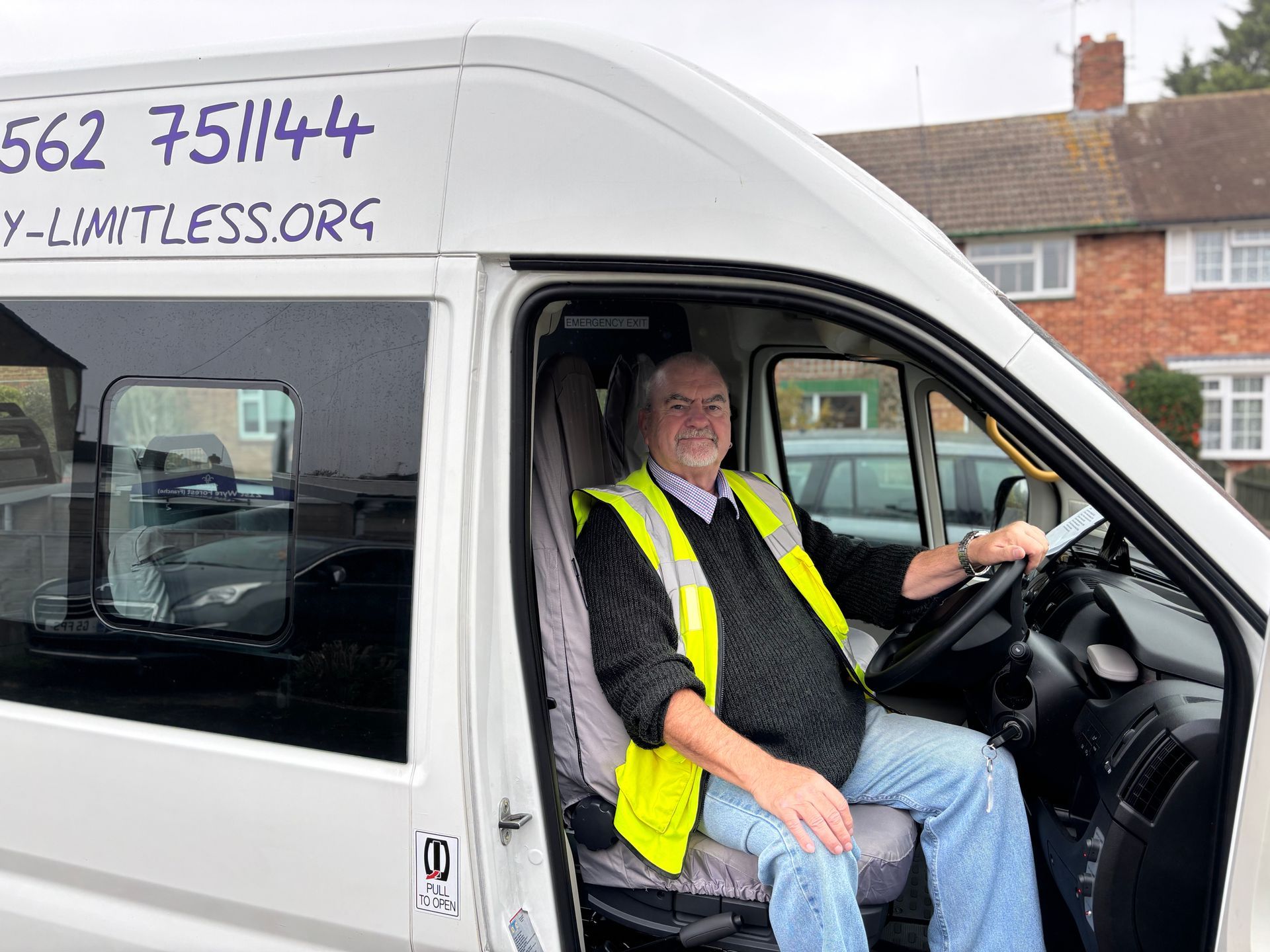 Man in yellow vest sits in driver's seat of white minibus smiling. Van has Simply Limitless website and phone number on side.