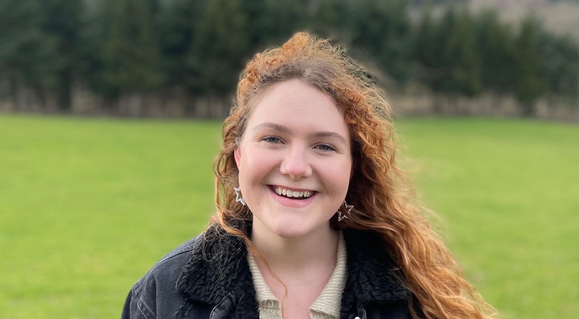 Woman with curly red hair smiles outside, wearing a black jacket and cream shirt; green field and trees in background.