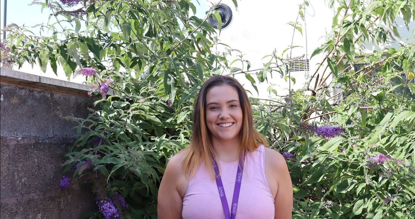 Woman in purple shirt smiles in front of green and purple foliage.
