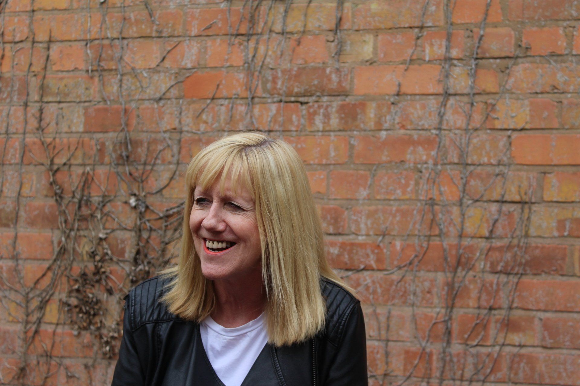 Woman with blonde hair smiles in front of a brick wall with vines. She wears a black jacket over a white shirt.