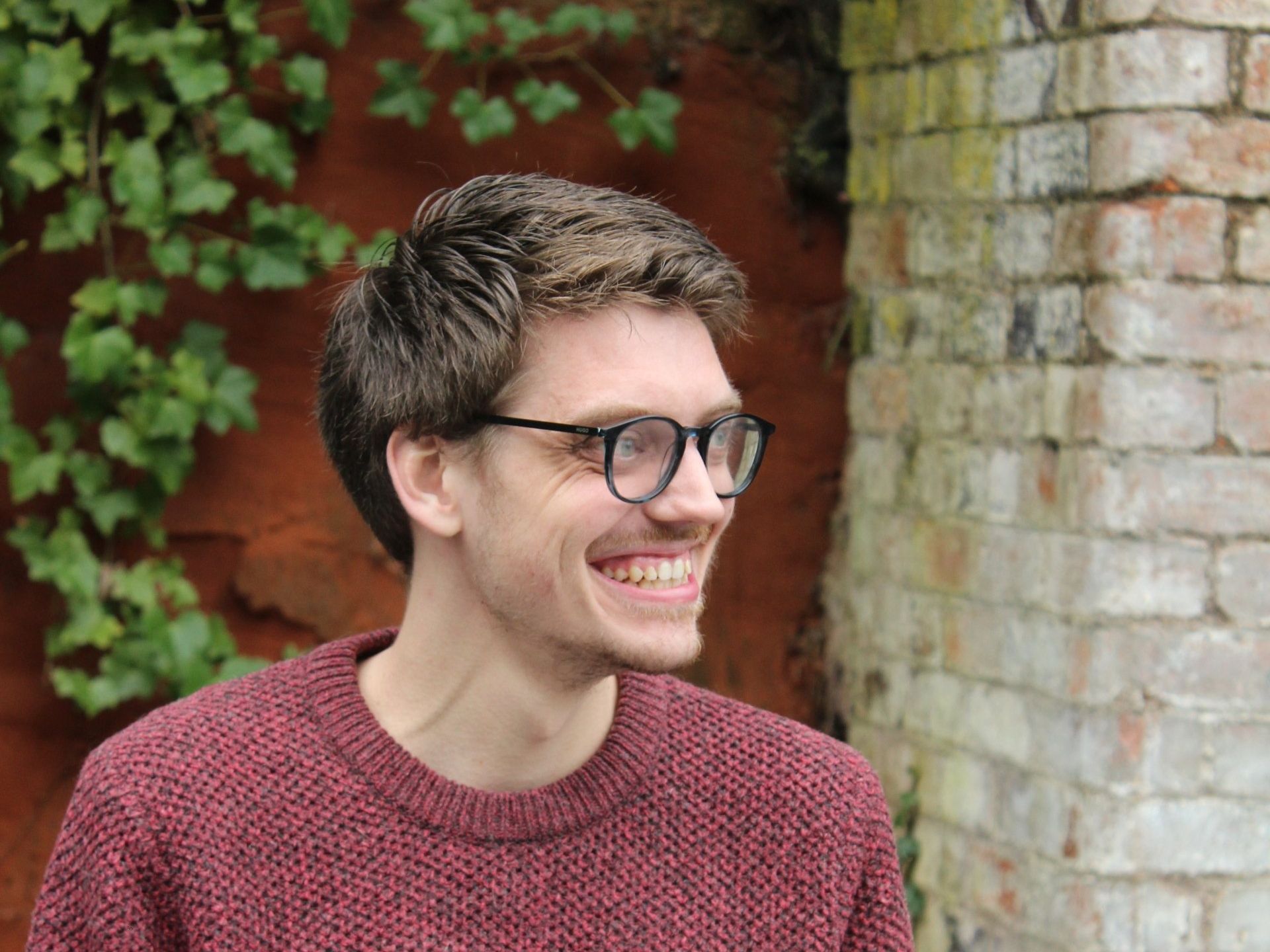 Smiling person in glasses and red sweater, next to a brick wall with ivy.