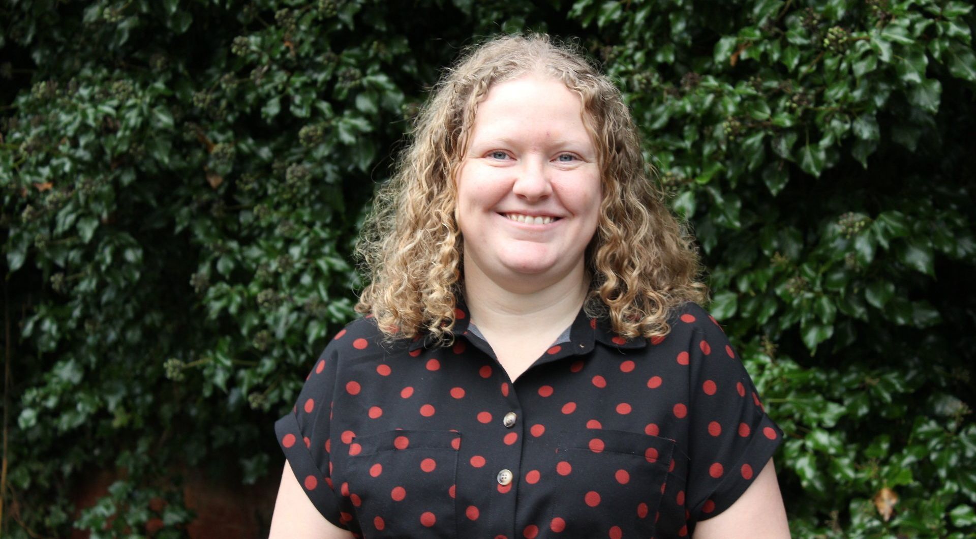Smiling person in glasses and red sweater, next to a brick wall with ivy.