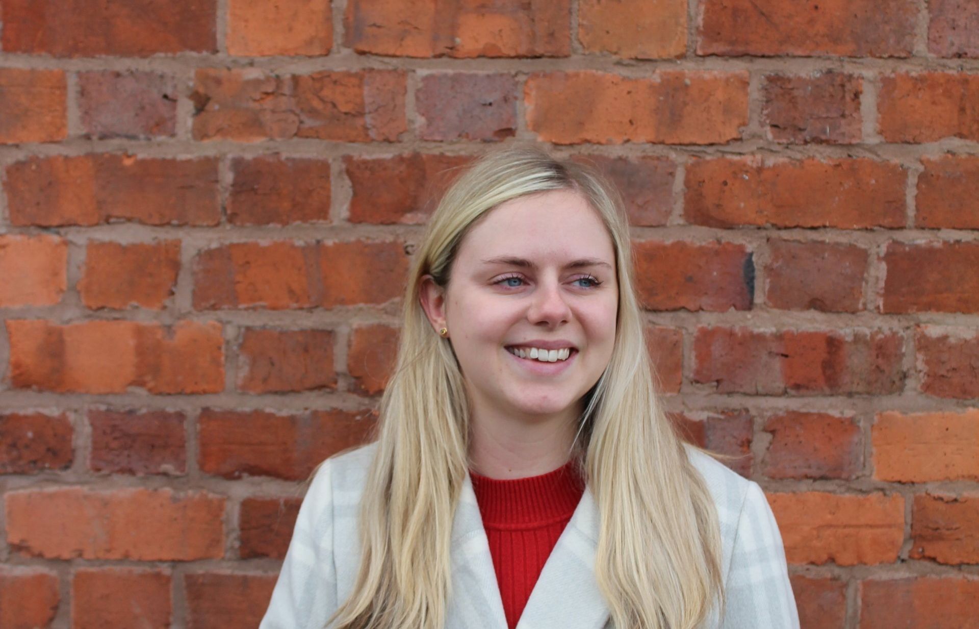 Woman with blonde hair smiles in front of a brick wall, wearing a red top and white coat.