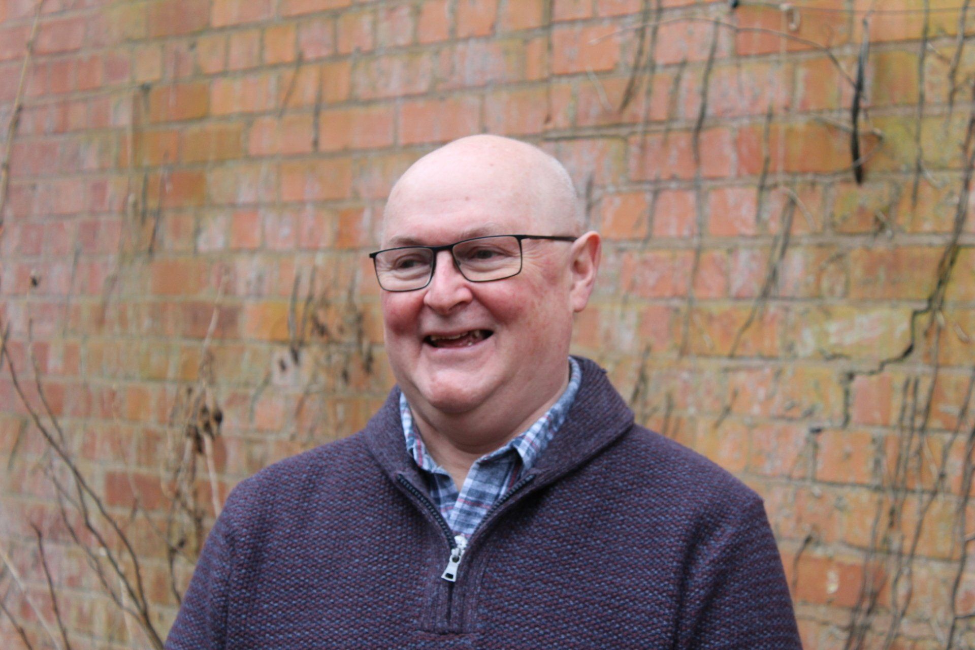 Man wearing glasses, smiling. He wears a blue plaid shirt under a dark purple sweater.