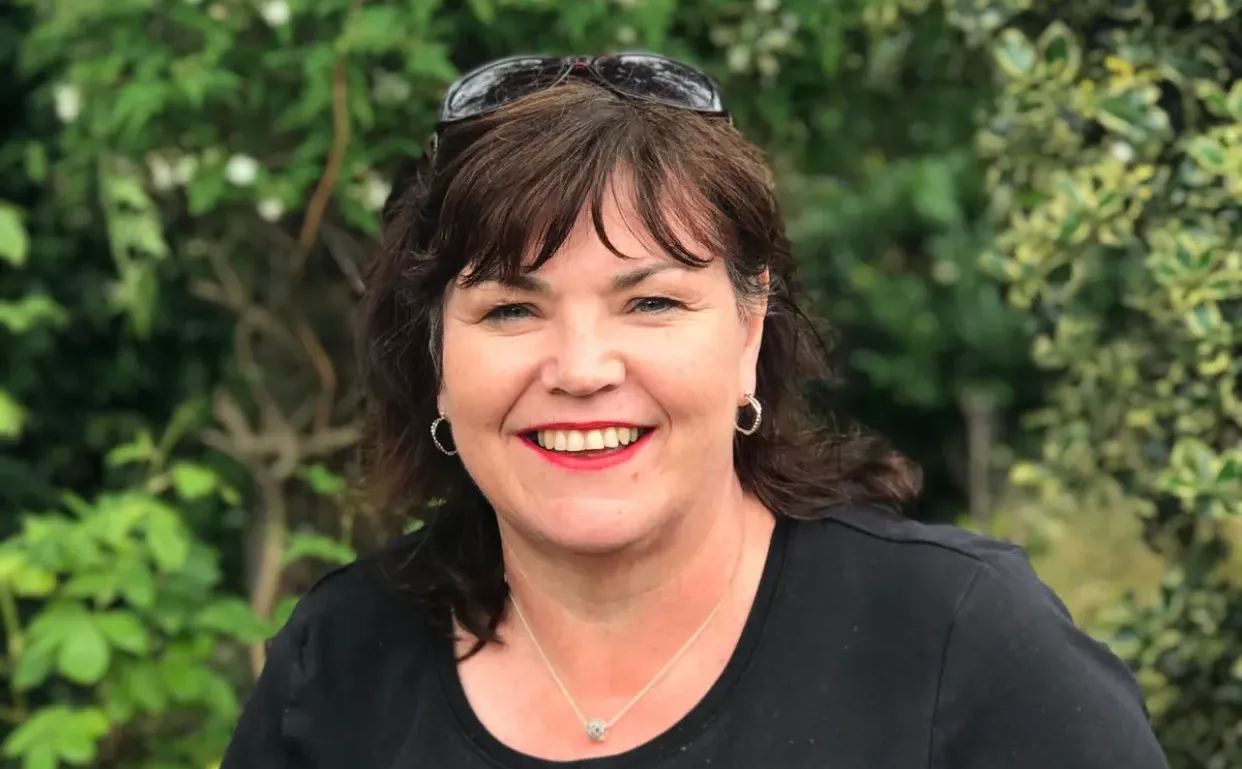 Woman with curly blonde hair smiling, wearing a black shirt with red polka dots, standing in front of green foliage.