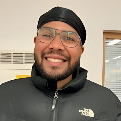 Person smiling, wearing glasses and a black durag, black jacket, white wall background.