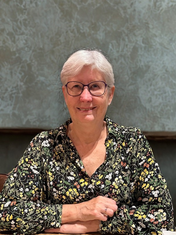 Woman with short white hair and glasses smiles. Wearing a floral shirt, she sits at a table.