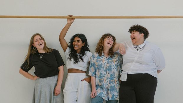 Four people laughing, posing together. One person holds a wooden bar above, smiling. White wall background.