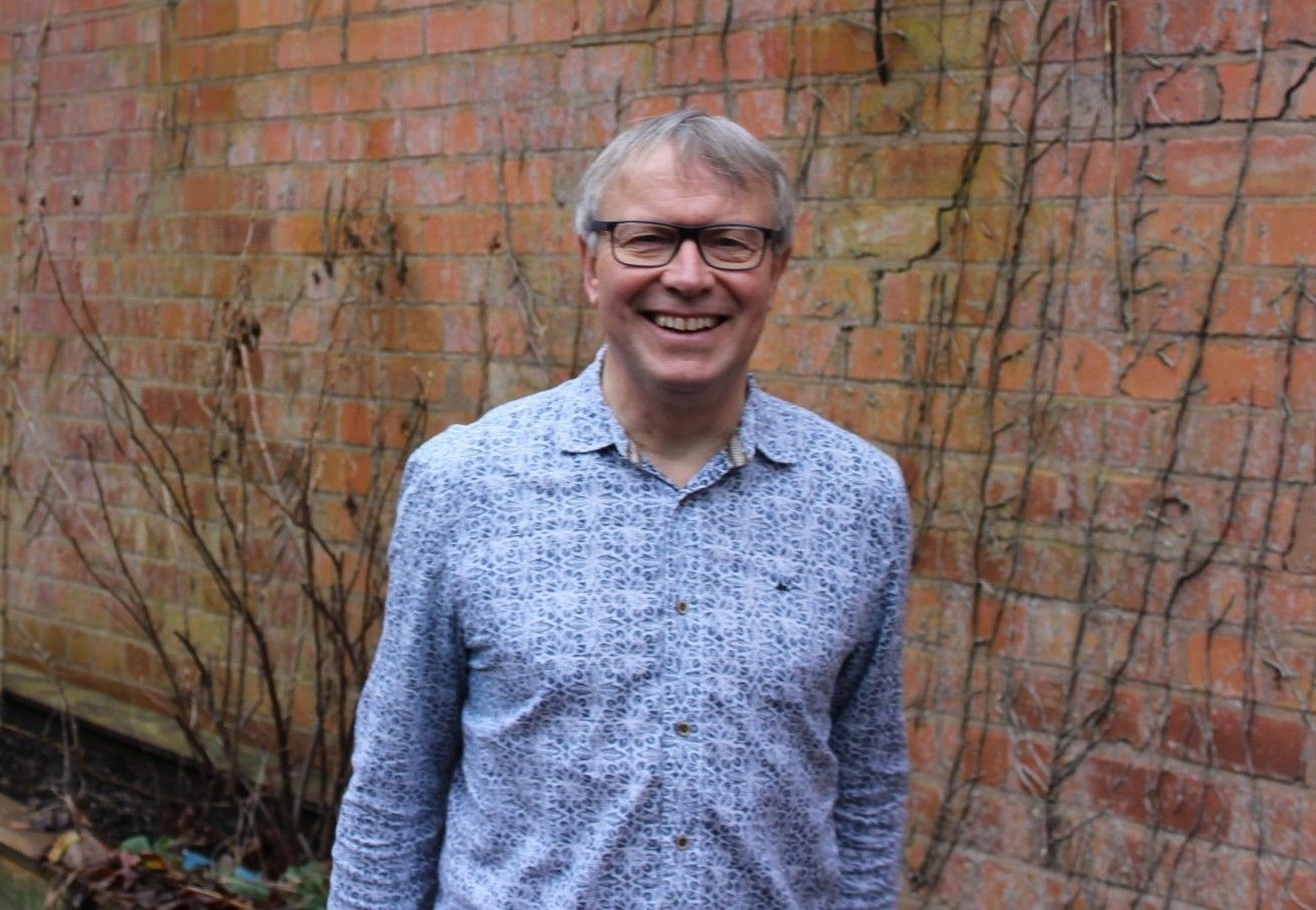 Woman with short white hair and glasses smiles. Wearing a floral shirt, she sits at a table.