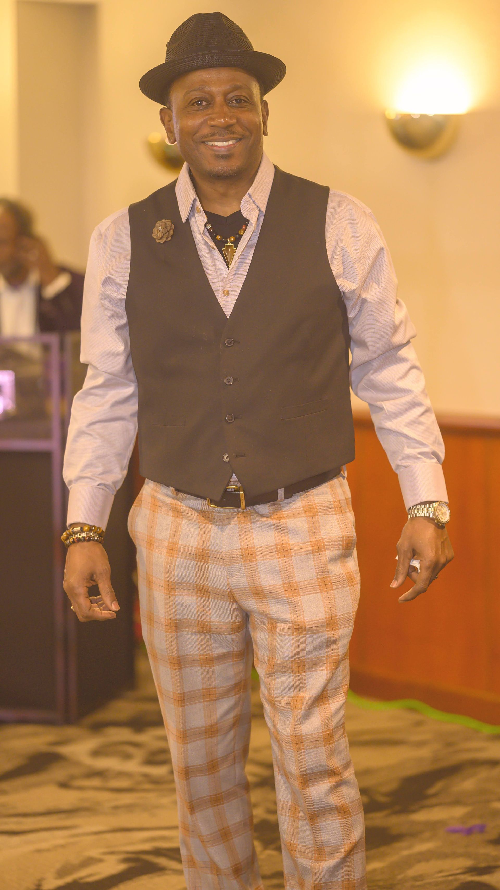 A man wearing a hat , vest and plaid pants is standing in a room.