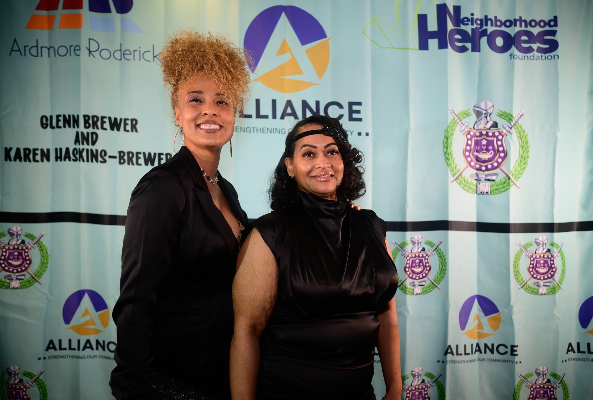 Two women are posing for a picture in front of a neighborhood heroes backdrop.