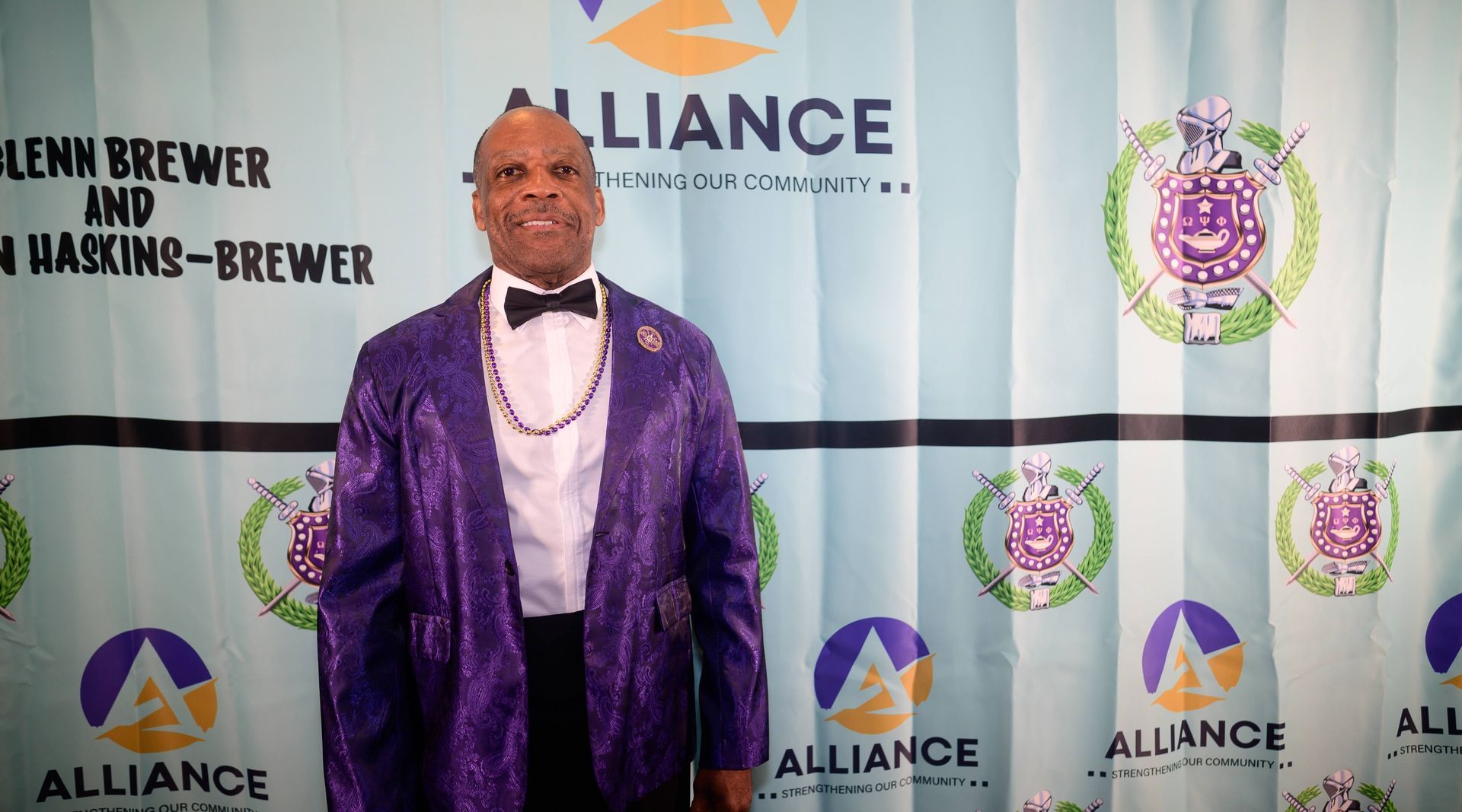 A man in a purple suit and bow tie is standing in front of a wall that says alliance