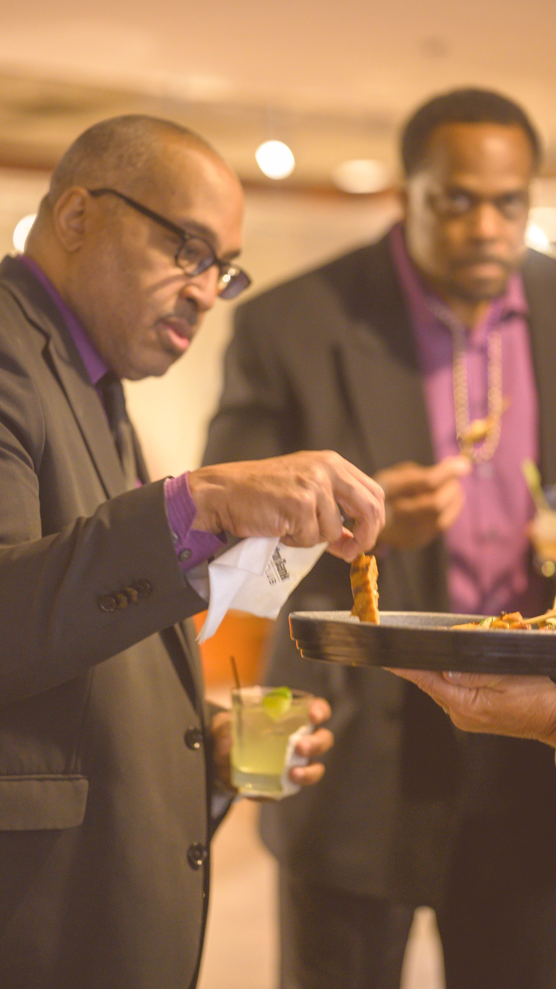 A man in a suit is serving food to another man in a purple shirt.