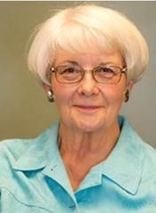 Elderly woman with short white hair, wearing glasses and a blue shirt, smiling.