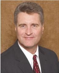 Man in suit and tie smiles at the camera. Brown background.