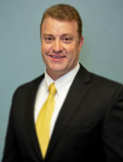 Man in a suit and yellow tie smiles in front of a teal background.