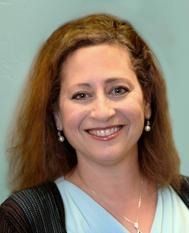 Woman with auburn hair, smiling, wearing a light blue shirt and pearl earrings.