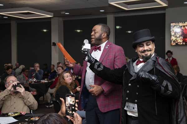 Man with microphone and magician in a room with people at tables; Man is speaking and magician smiles.
