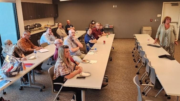 Group of people seated at tables, listening to a woman presenting in a meeting room.