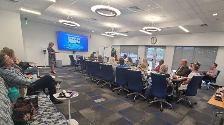 A person presents to a group seated at a long table in a modern conference room.