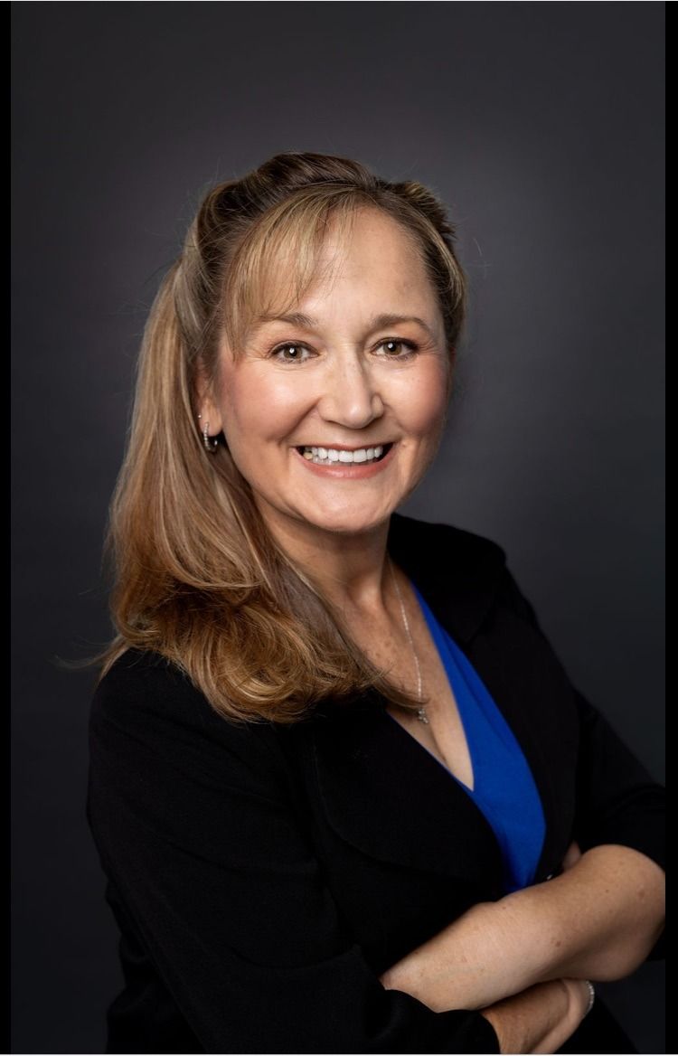 Woman with blonde hair smiles, arms crossed, wearing a black blazer over a blue top; gray background.