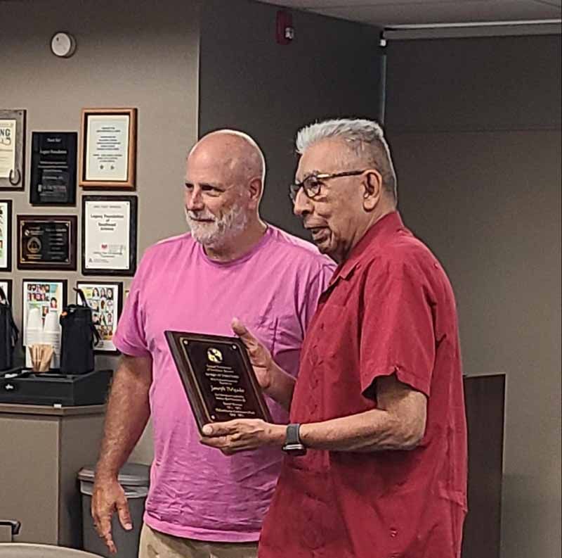 Two men hold a plaque. Older man in red shirt presents it to a bald man in pink shirt. Office setting.