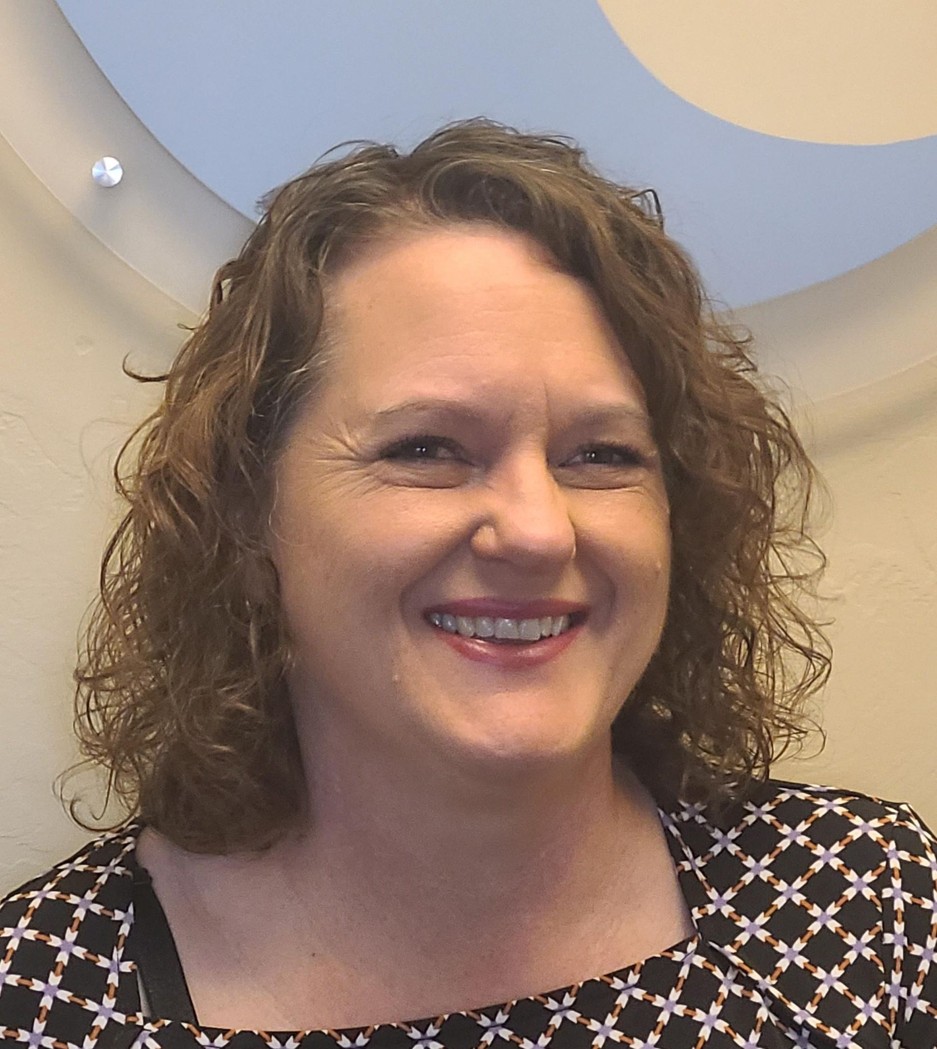 Woman with curly brown hair smiles, wearing a black and white patterned top, indoors.
