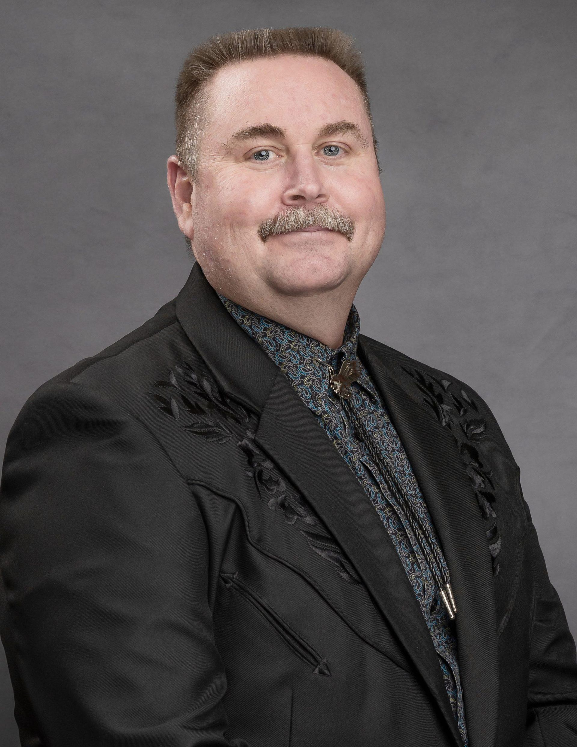 Man in a black blazer with embroidery, bolo tie, and mustache, looking at the camera.