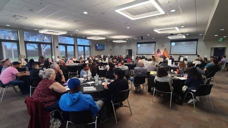 Large conference room with people seated at tables. Two screens display presentations.