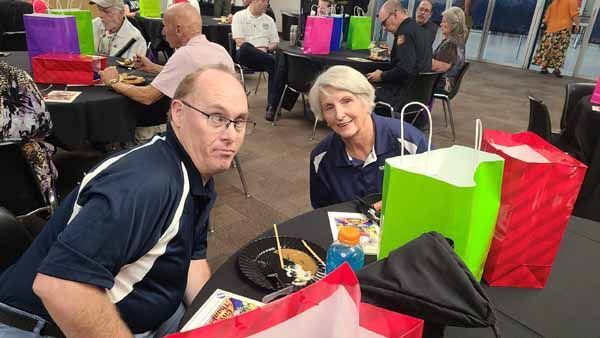 A couple at a table with gifts, smiling. Other people sit at tables in a room.