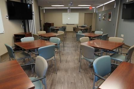 A well-lit classroom with tables and chairs arranged for seating. There is a whiteboard at the far end of the room.