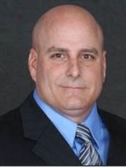 Bald man in suit and tie, smiling slightly. Blue shirt, dark suit jacket.