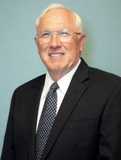 White-haired man in a suit and tie smiles, wearing glasses, against a blue-green background.