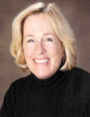 Blonde woman smiles, wearing a black turtleneck, against a blurred background.