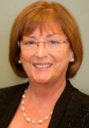 Woman with short red hair, glasses, and pearl necklace smiles at camera.